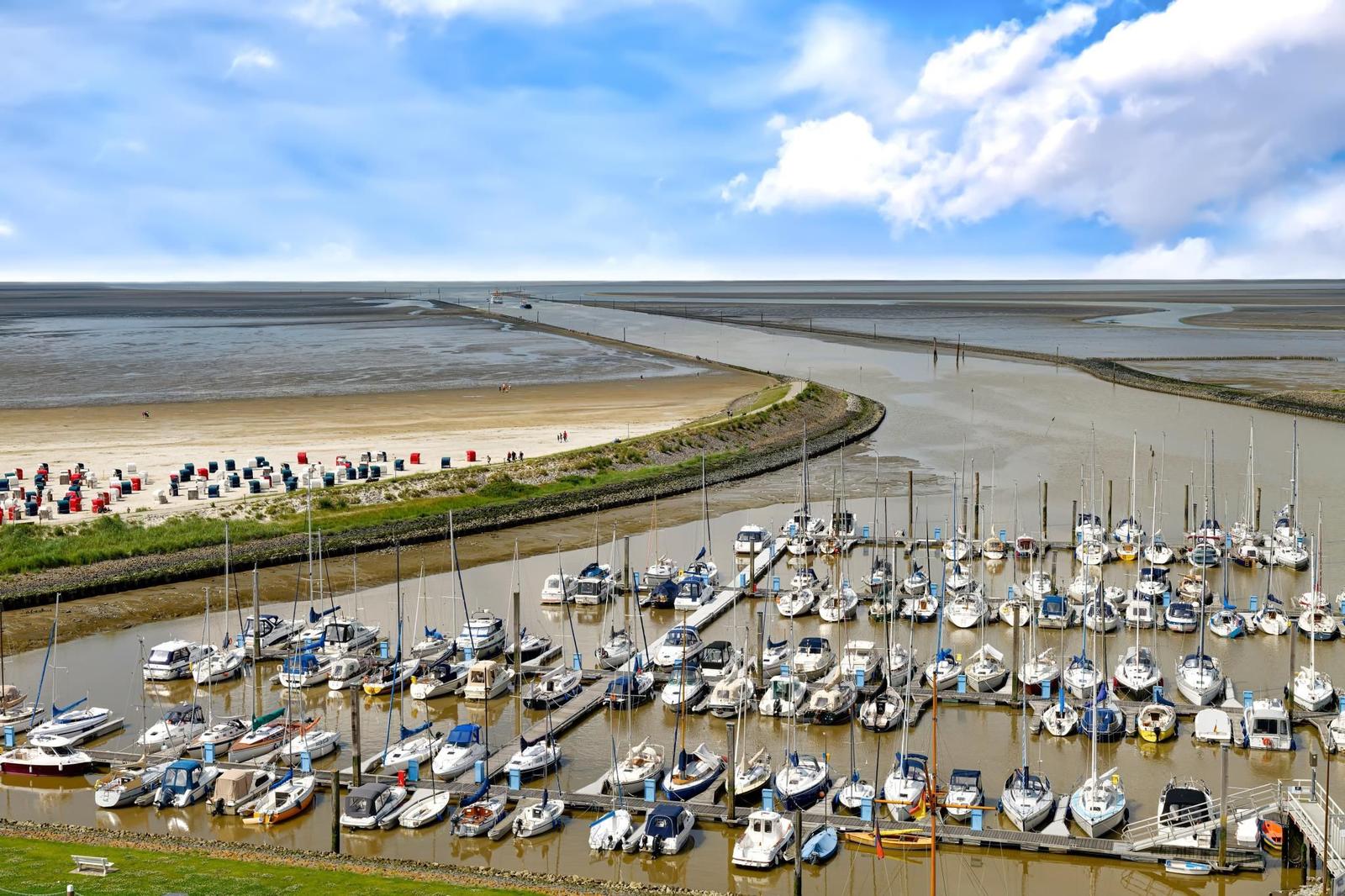 Hafen mit vielen Booten, Strand und Wattgebiet unter blauem Himmel.