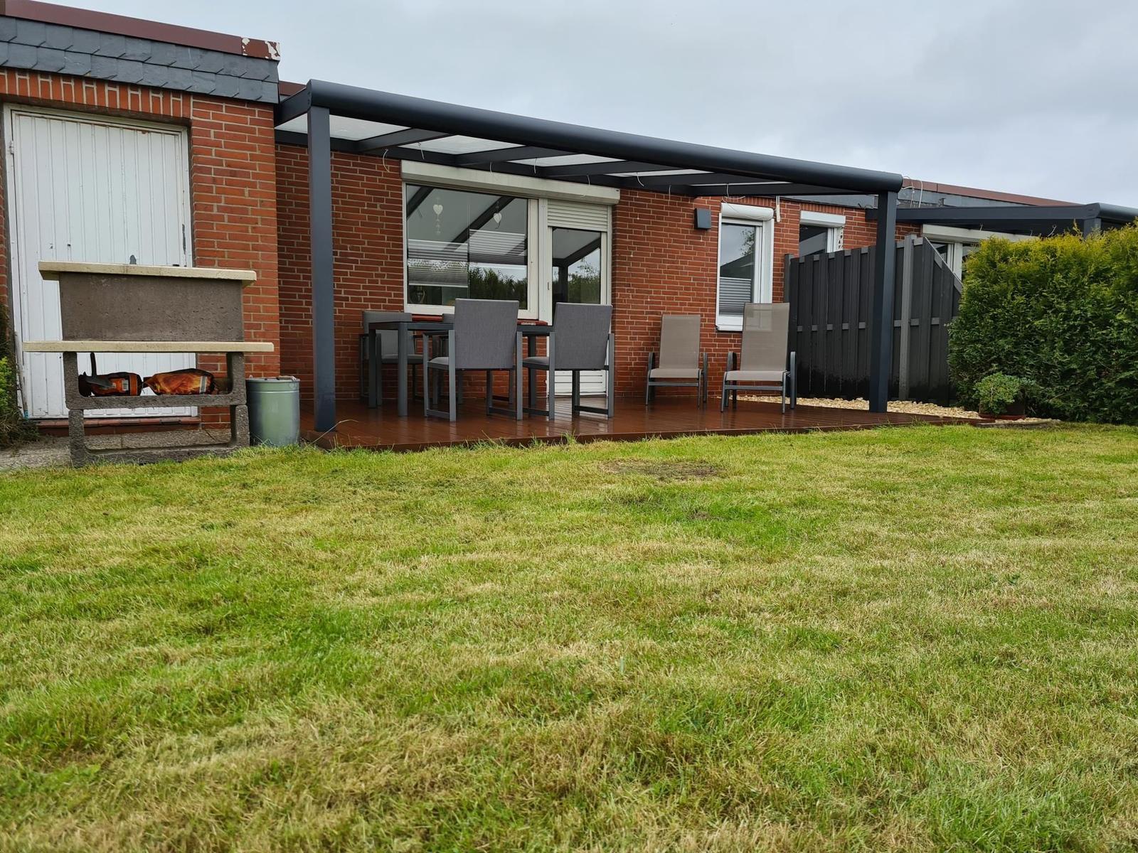 Terrasse mit Tisch, Stühlen und Sitzgelegenheiten. Grasfläche und Grillbereich vor Haus.