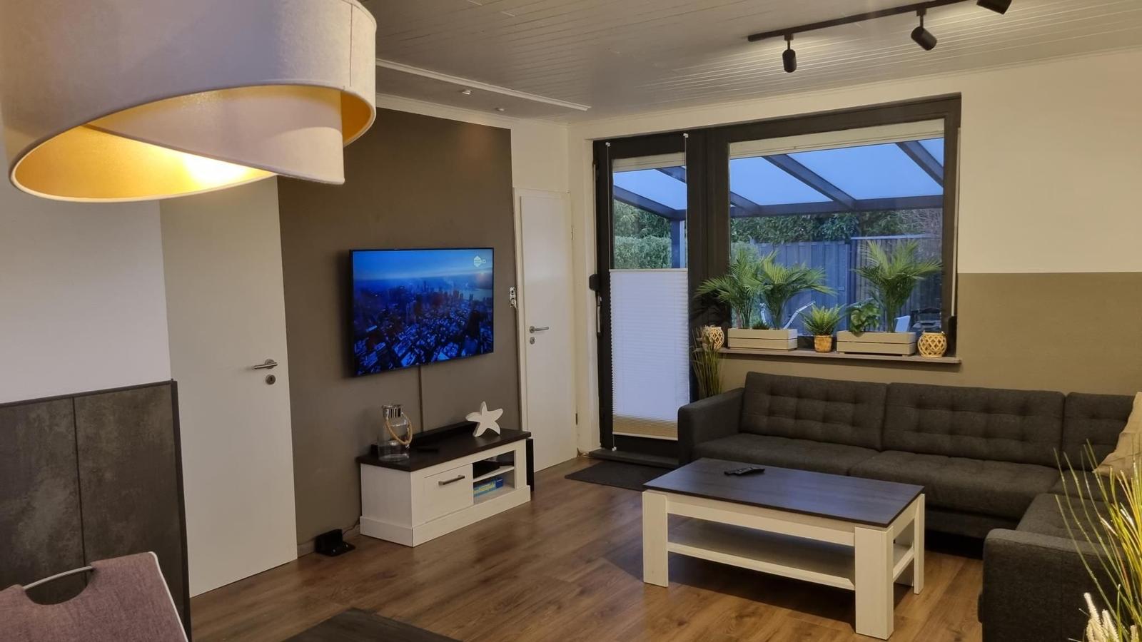 Wohnzimmer mit Sofa, Couchtisch und Fernseher. Große Fenster mit Blick auf Terrasse.