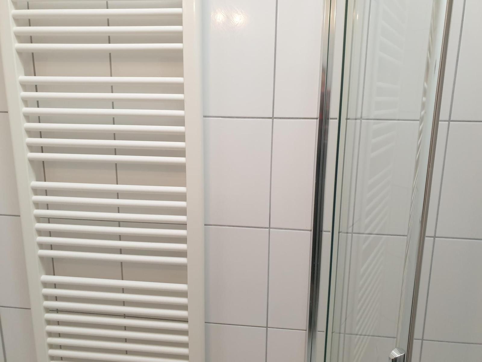 Shower area with glass door and heater against white tiled wall
