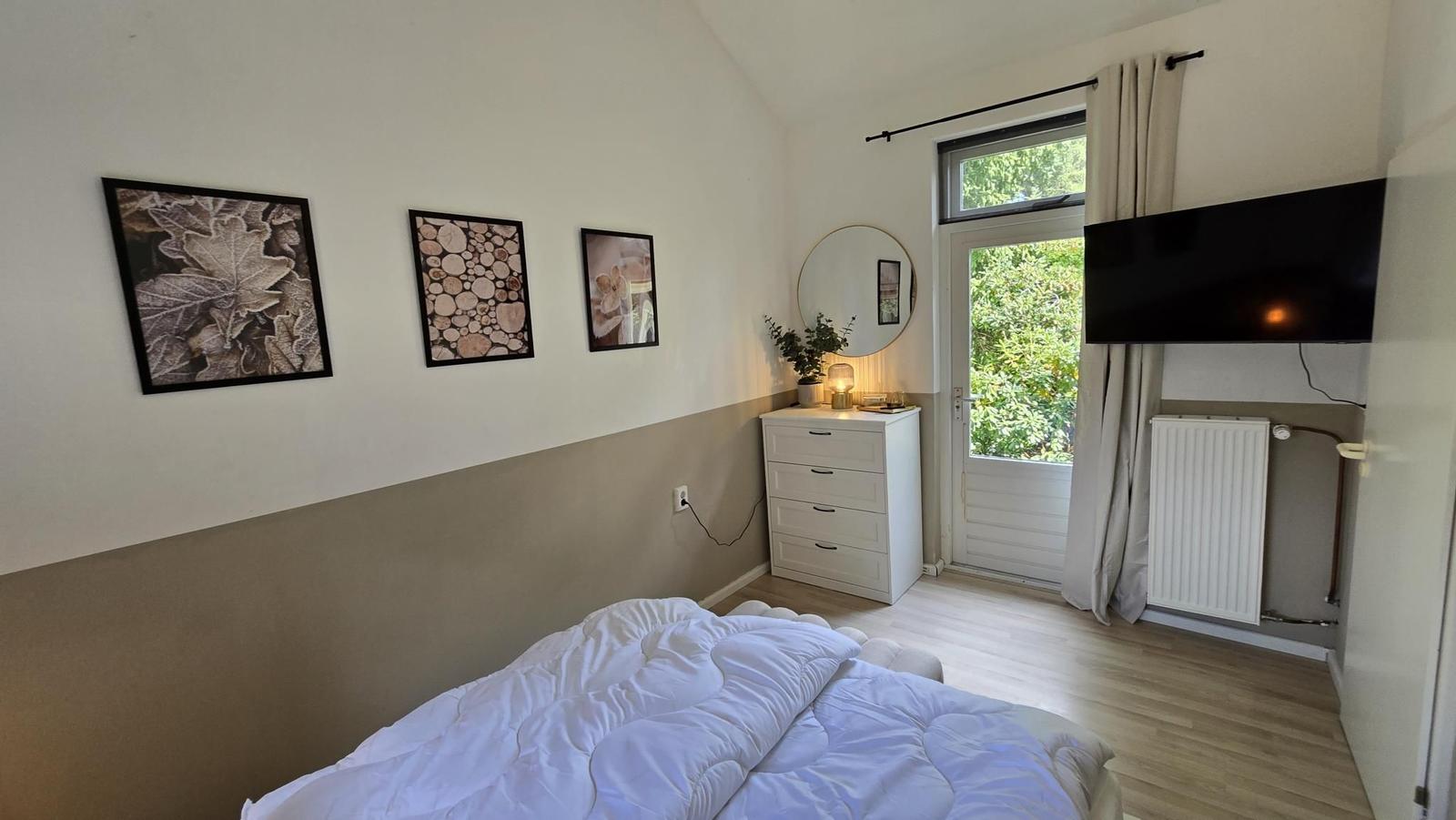 Schlafzimmer mit Bett, Schrank, Fernseher und Fenster zur grünen Außenseite.