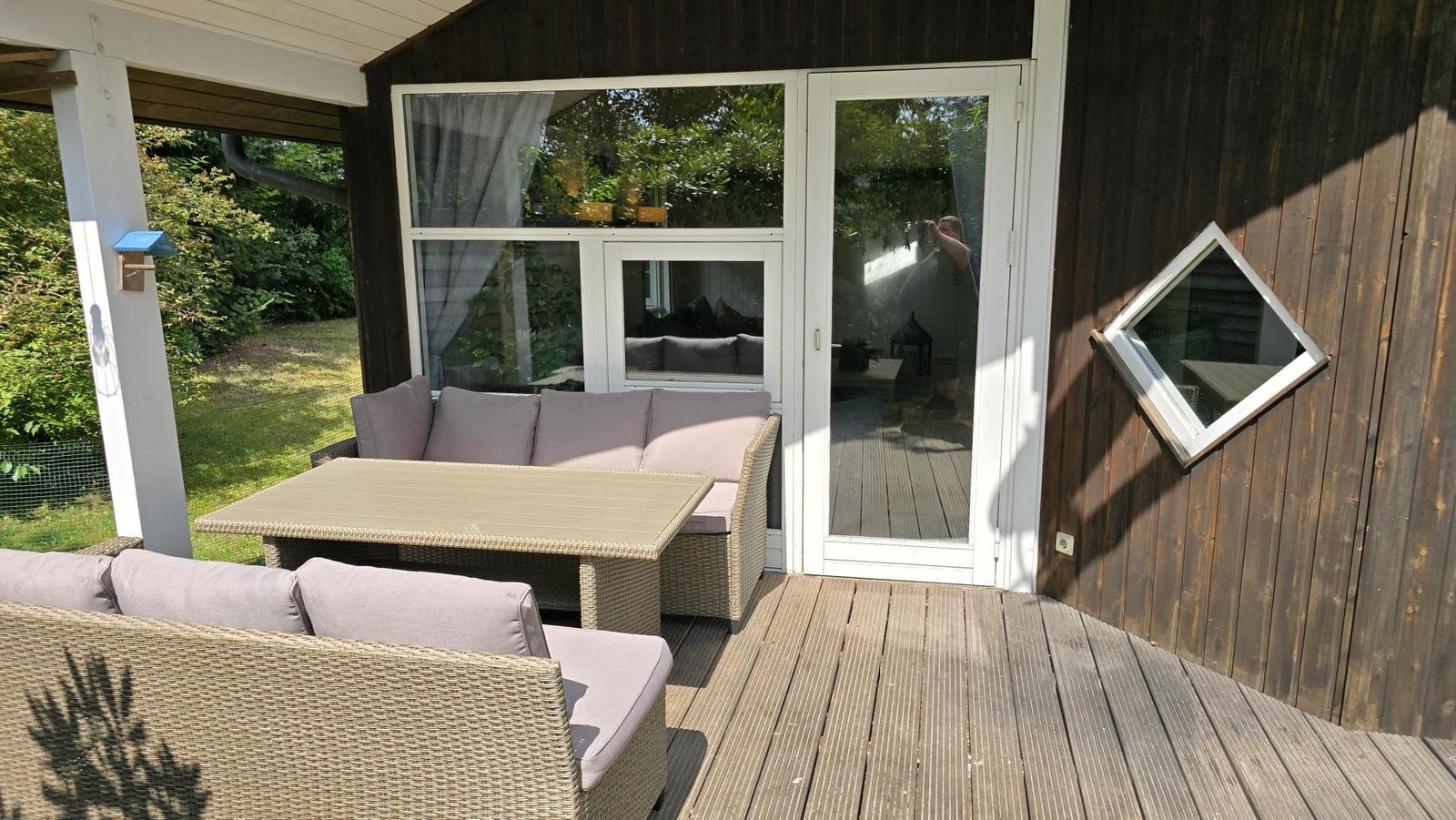 Terrasse mit Sitzgruppe und Tisch. Holzwand und Glastür zum Innenraum.