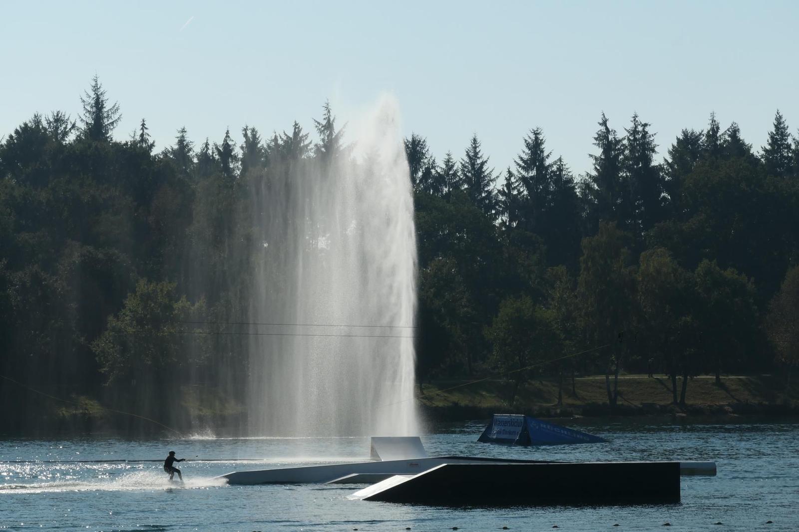 Wasserskifahrer beim Springen über Sprungbrett und Fontäne am See.