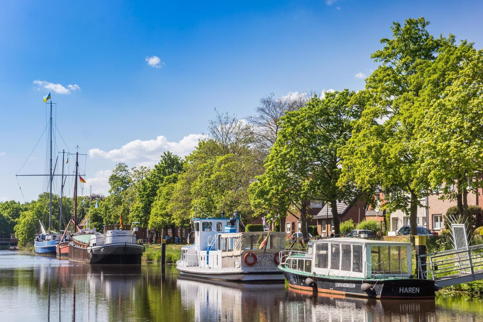 Schiffe liegen am Kanal, umgeben von Bäumen und Häusern unter blauem Himmel.