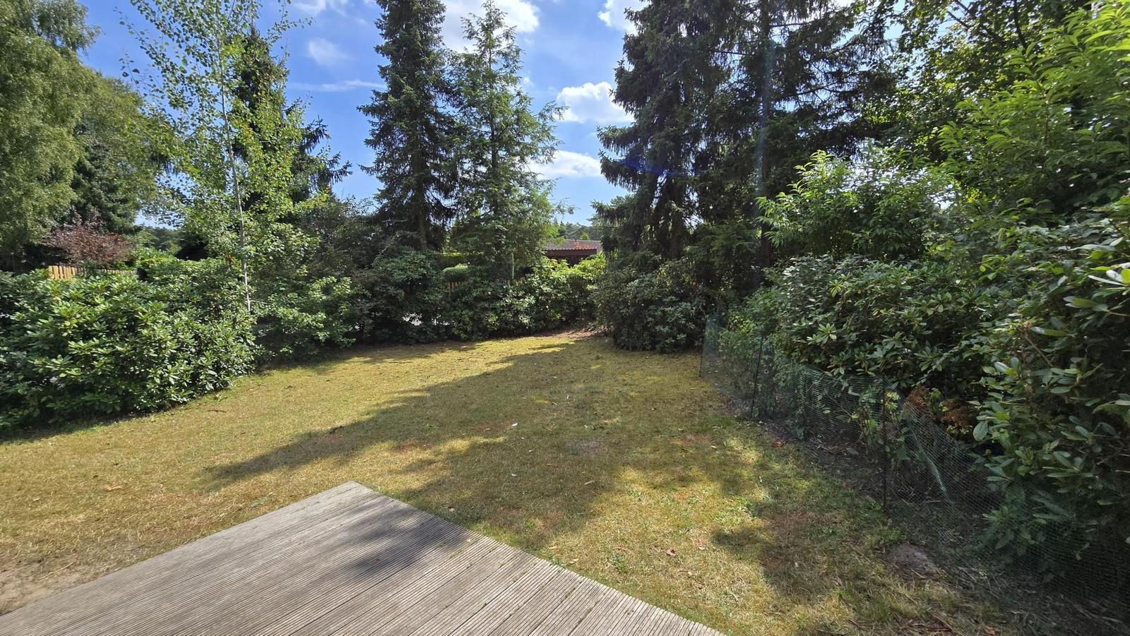 Grüner Garten mit Holzterrasse und Bäumen im Hintergrund.