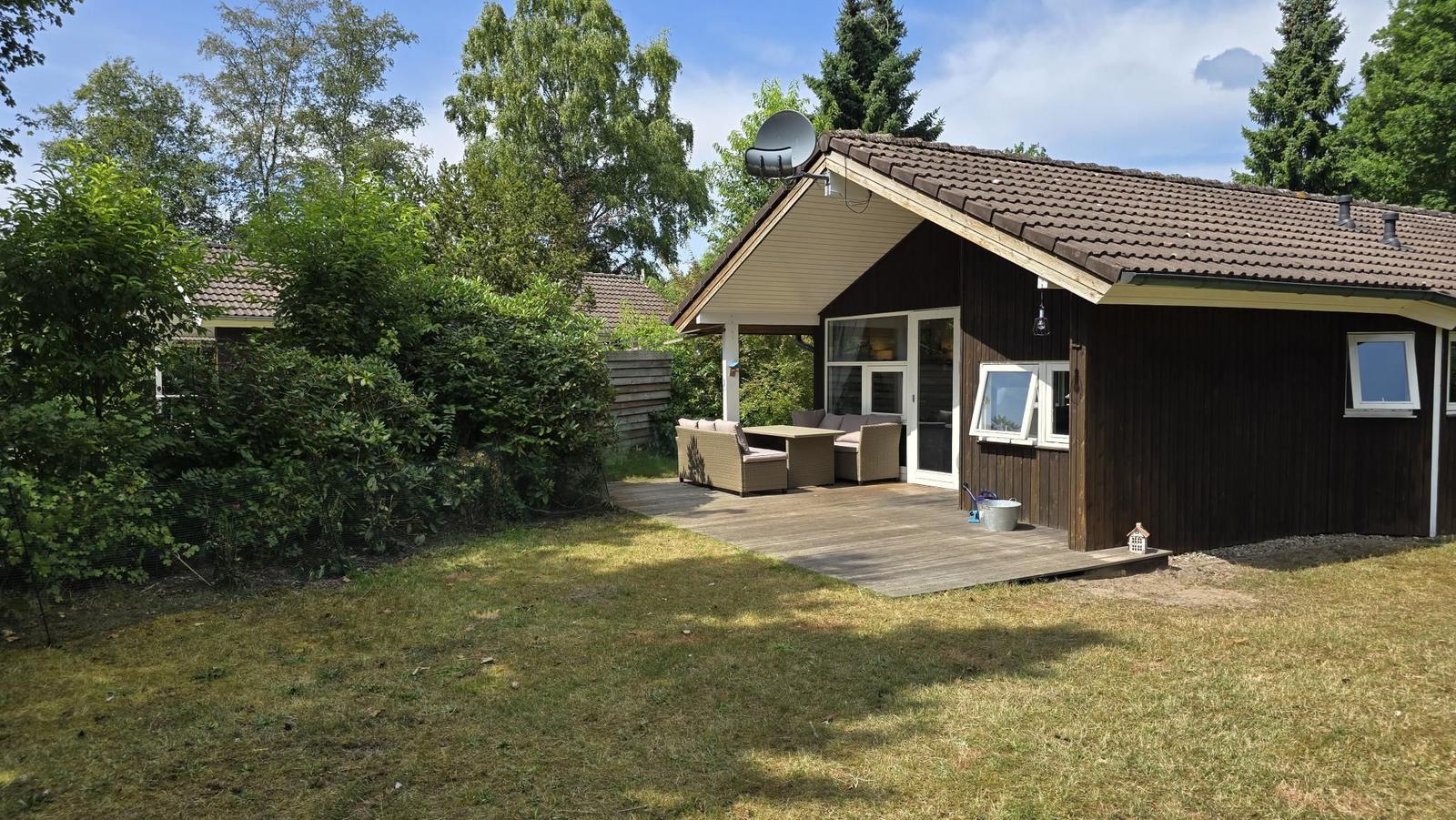 Holzhaus mit Terrasse, Garten und Satellitenanlage. Gartenmöbel und Fenster sichtbar.