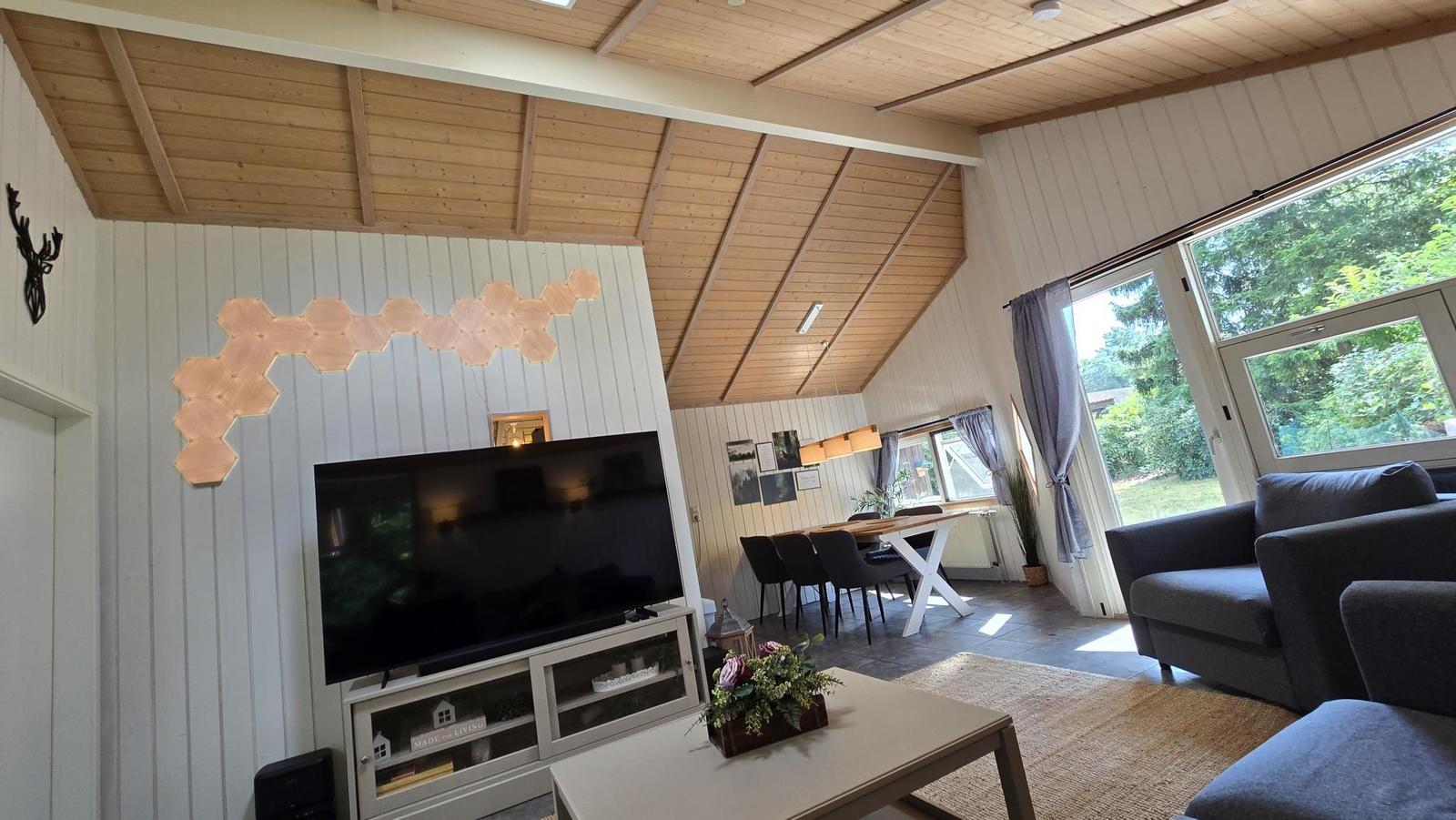 Wohn- und Essbereich mit TV, Sofa und großem Fenster. Holzdecke und weiße Wände.