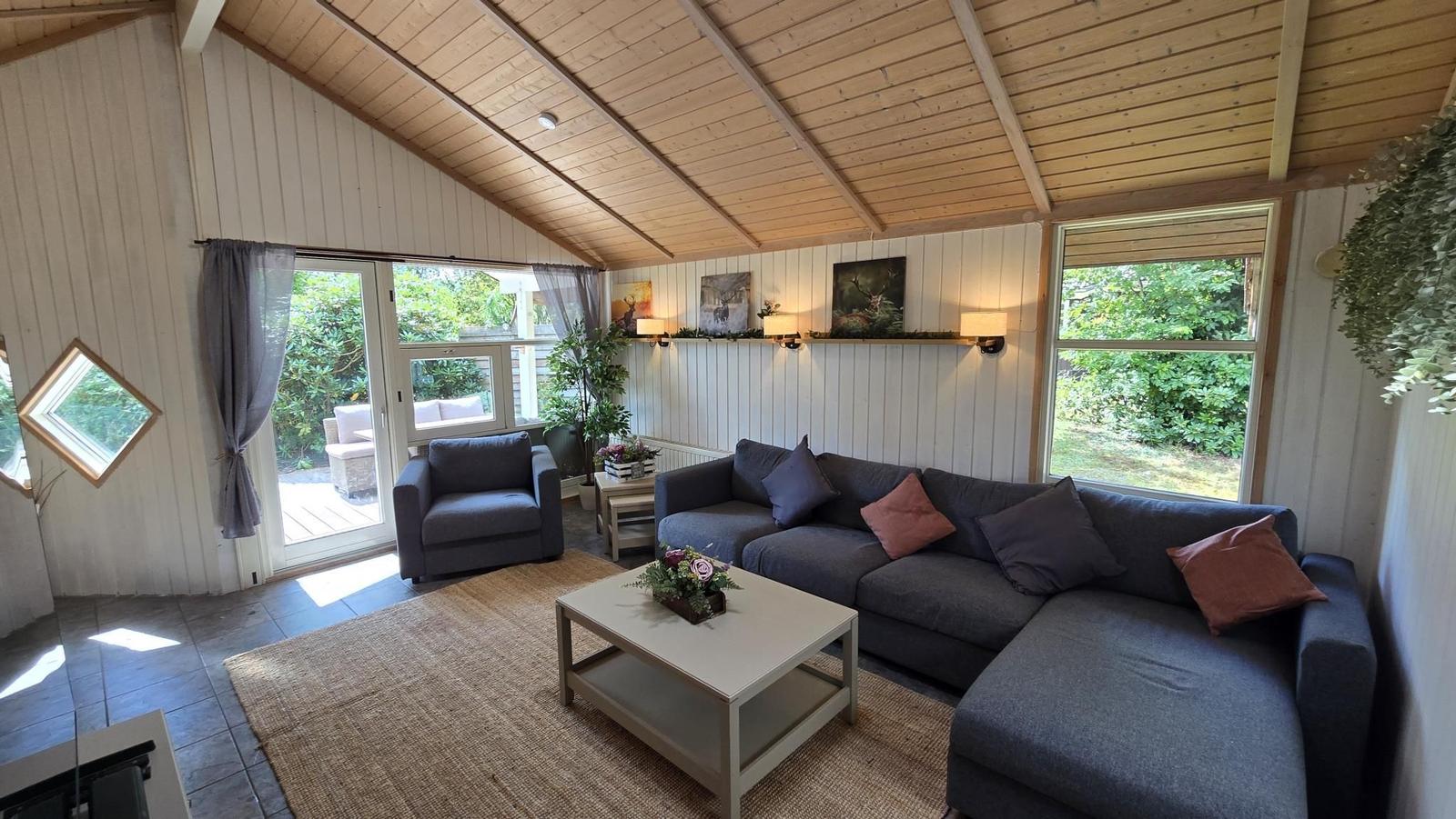 Wohnzimmer mit Sofa, Sessel und Tisch. Holzdecke und Fenster zur Außenterrasse.