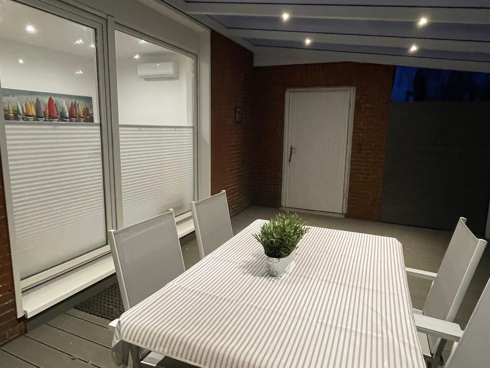 Terrasse mit Tisch, Stühlen und Pflanze. Große Fenster und rote Wand.