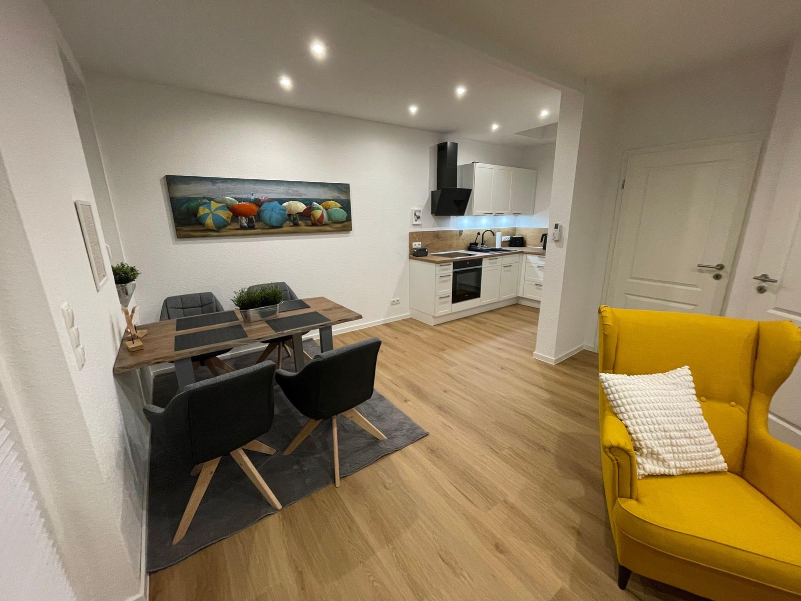 Essbereich mit Tisch, Stühlen und Wandbild. Küche mit Arbeitsplatte und Ofen. Gelber Sessel mit Kissen.