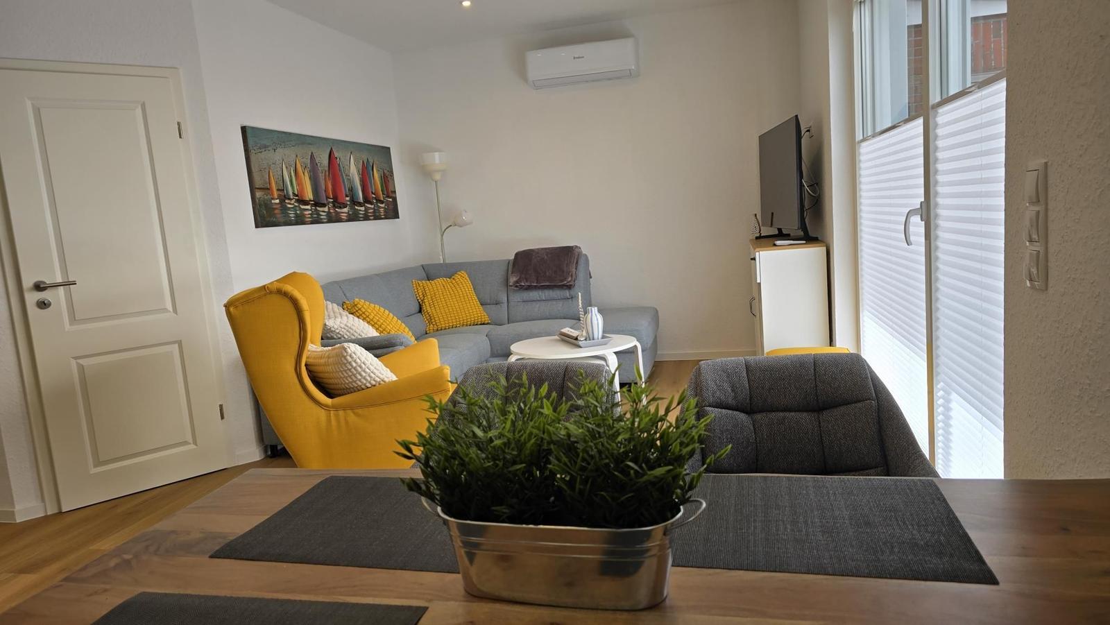 Wohnzimmer mit Sofa, gelber Sessel und TV. Fenster mit Jalousien.