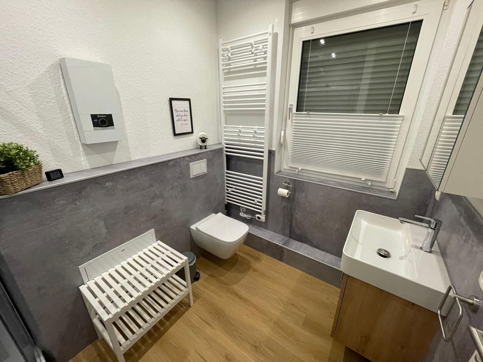 Badezimmer mit Toilette, Waschbecken und Heizung. Holzboden und Fenster mit Jalousie.