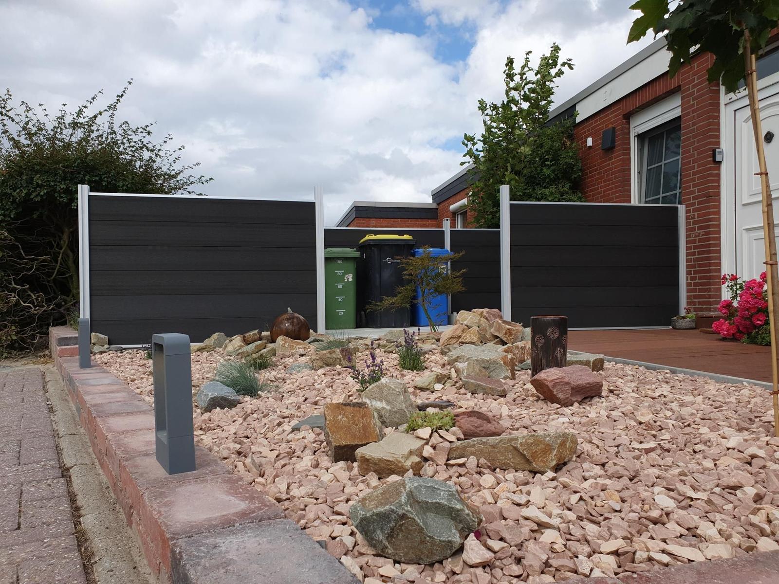 Hinterhof mit Steinbeete, dunklem Zaun und Terrasse. Mehrere Mülltonnen sind sichtbar.