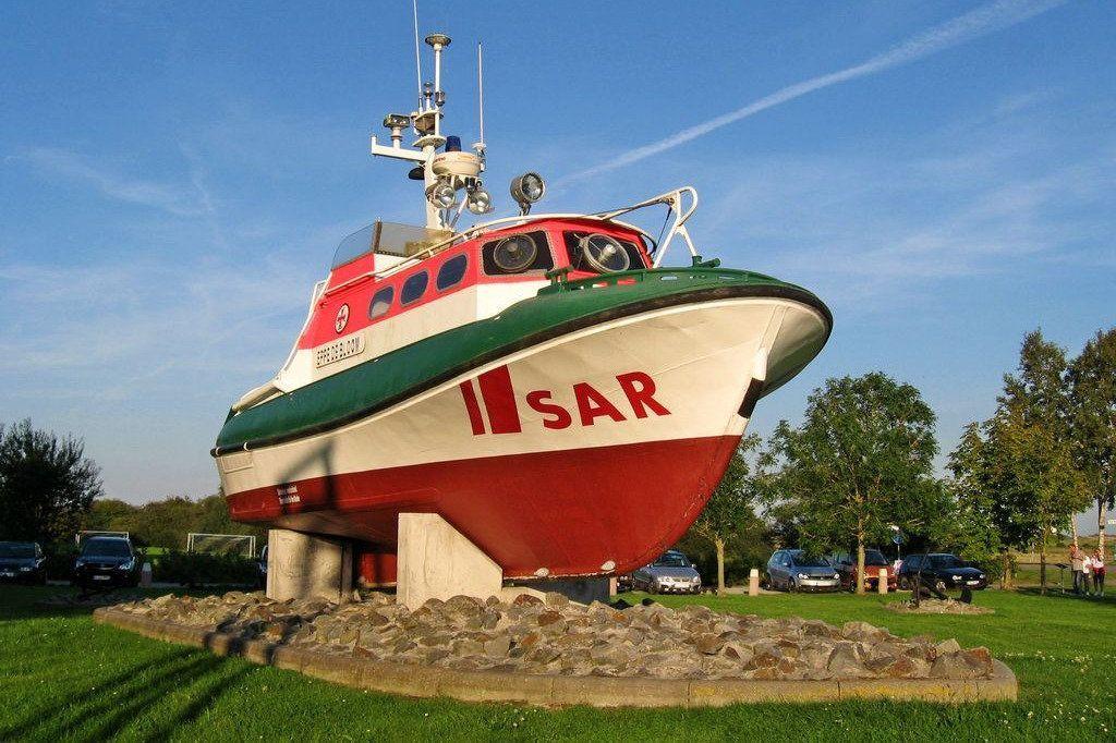 SAR-Schiff als Skulptur auf grünem Rasen mit Steinen und Bäumen im Hintergrund.