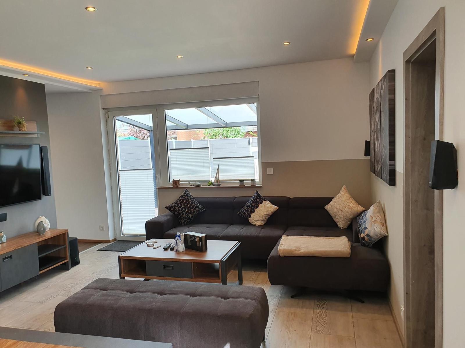 Wohnzimmer mit Sofa, Couchtisch und TV-Wand. Fenster mit Blick auf Garten.