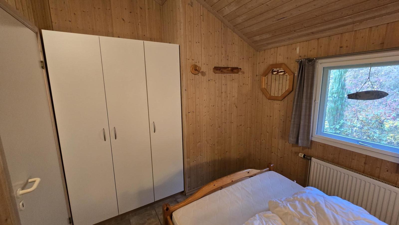 Schlafzimmer mit Holzwand, Bett, Kleiderschrank und Fenster mit Blick in den Wald.