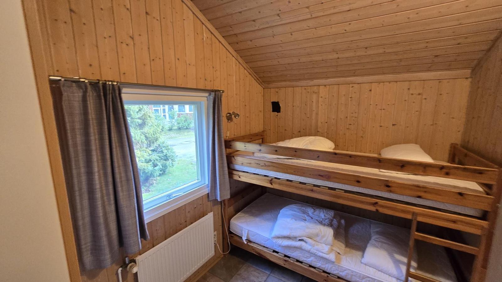 Zimmer mit Holzwand und Doppelstockbett. Fenster mit Vorhang und Heizkörper.