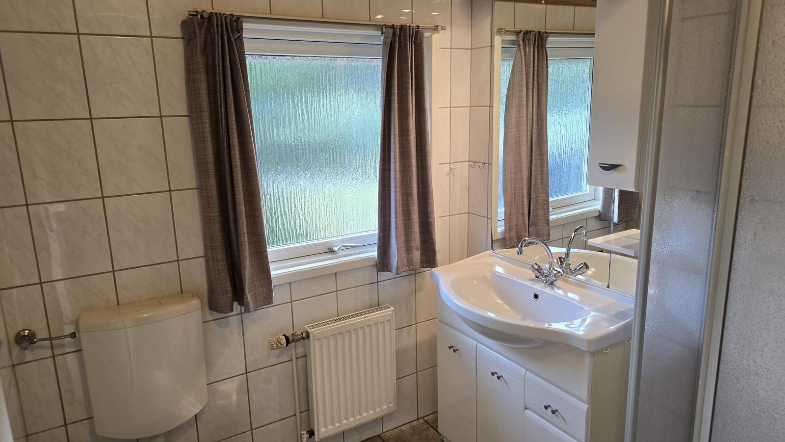 Badezimmer mit Waschbecken, Spiegel, Toilette und Fenster mit Vorhängen.