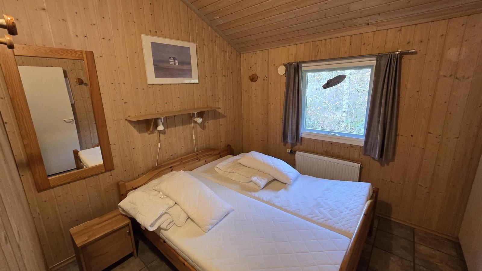 Schlafzimmer mit Holzwand, Bett, Spiegel und Fenster mit Blick auf Wald.