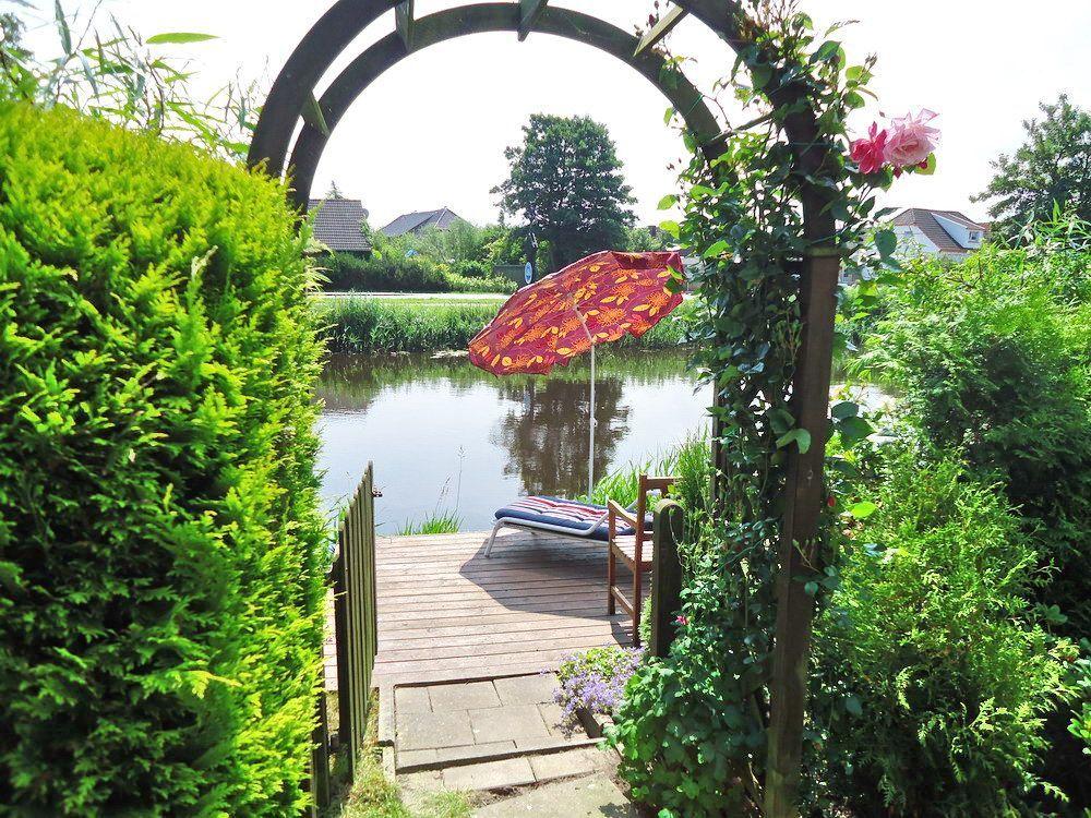 Durch einen Bogen führt ein Weg zu einer Terrasse mit Liege und Sonnenschirm am Wasser.