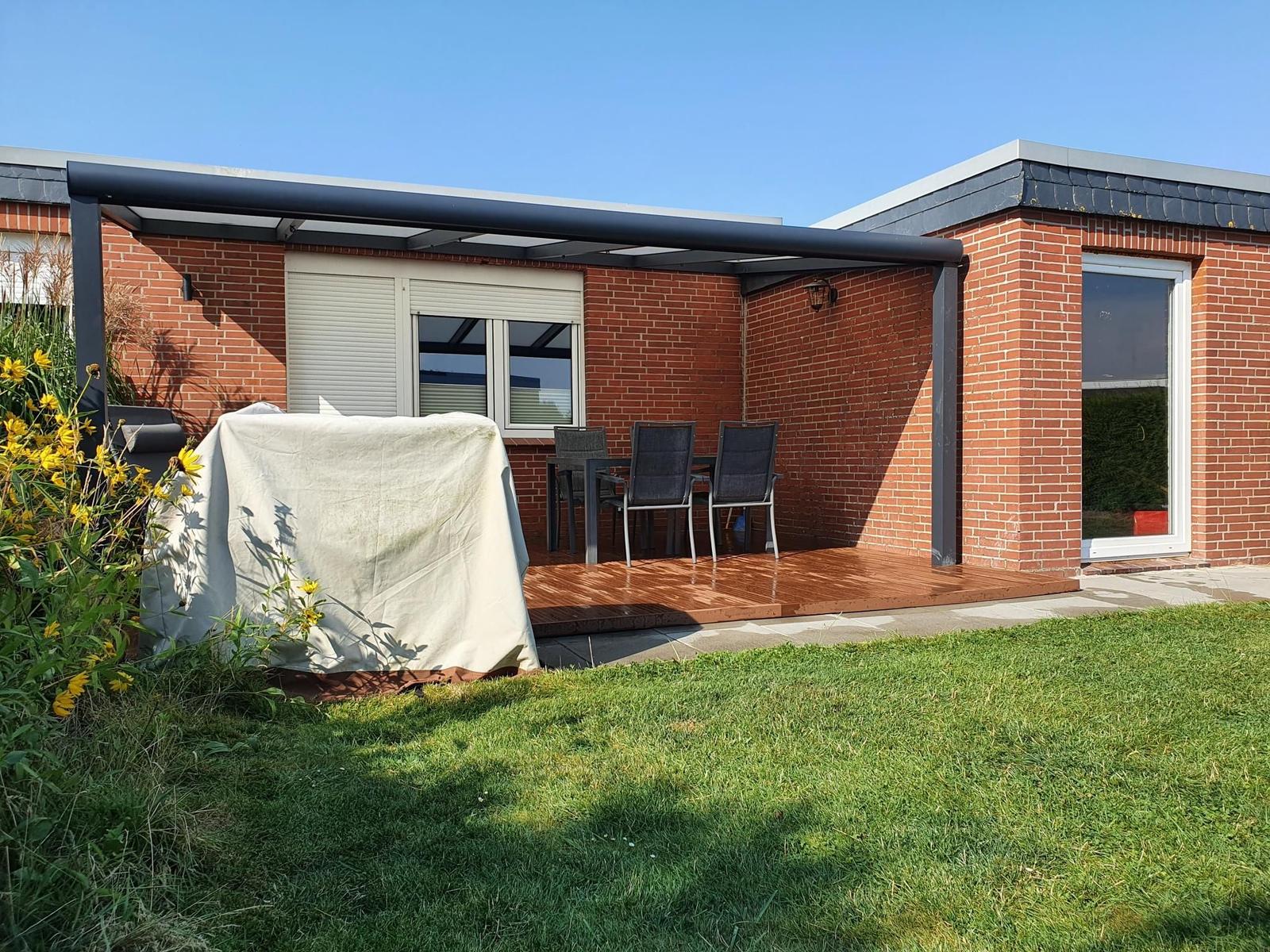 Terrasse mit Tisch, Stühlen und Grill unter Pavillon. Rasen und Blumen im Vordergrund.