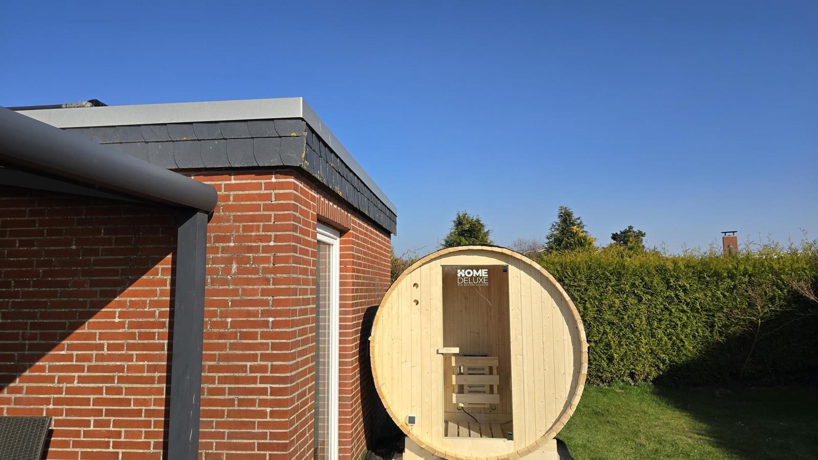 Außenliegende Holzsauna neben einem Backsteinhaus mit grünem Rasen und Hecken.