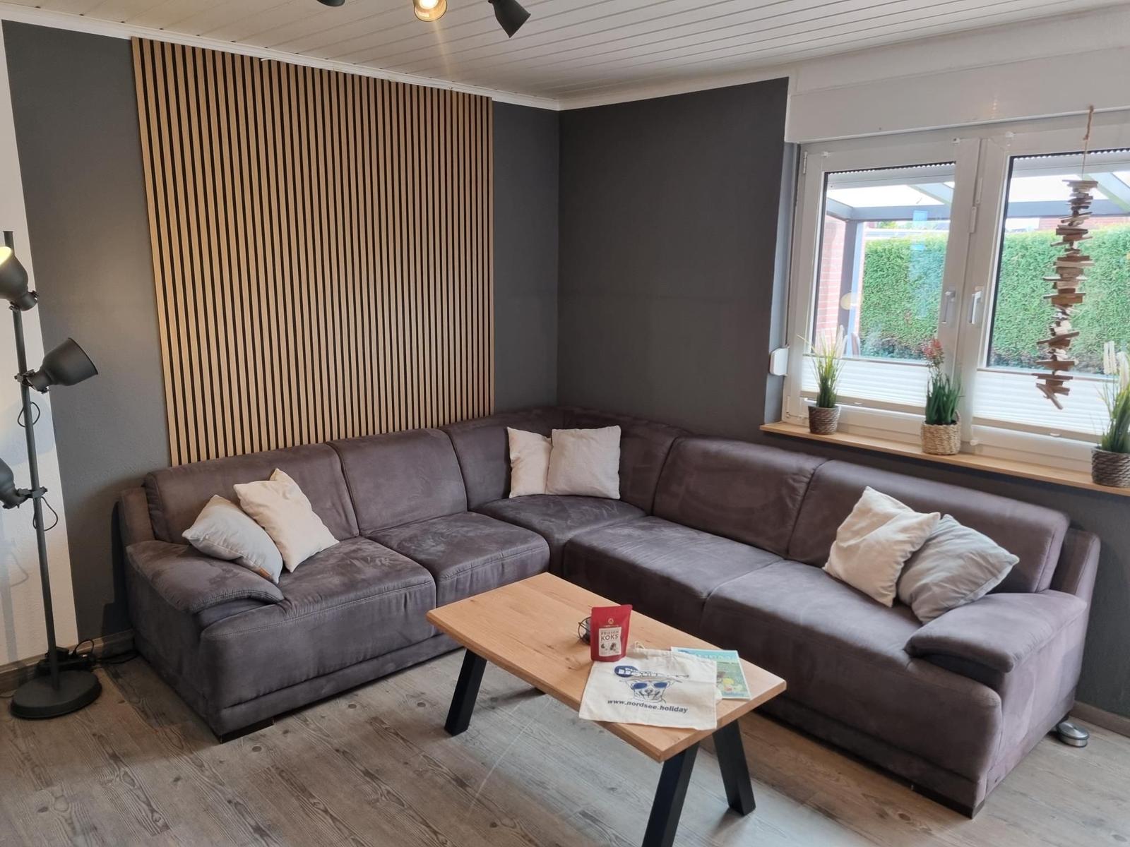 Ein Wohnzimmer mit grauem Sofa, Holztisch und Fenster mit Pflanzen.