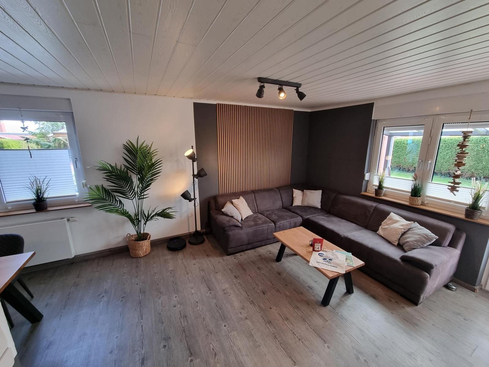 Wohnzimmer mit Ecksofa, Holzboden und großen Fenstern.