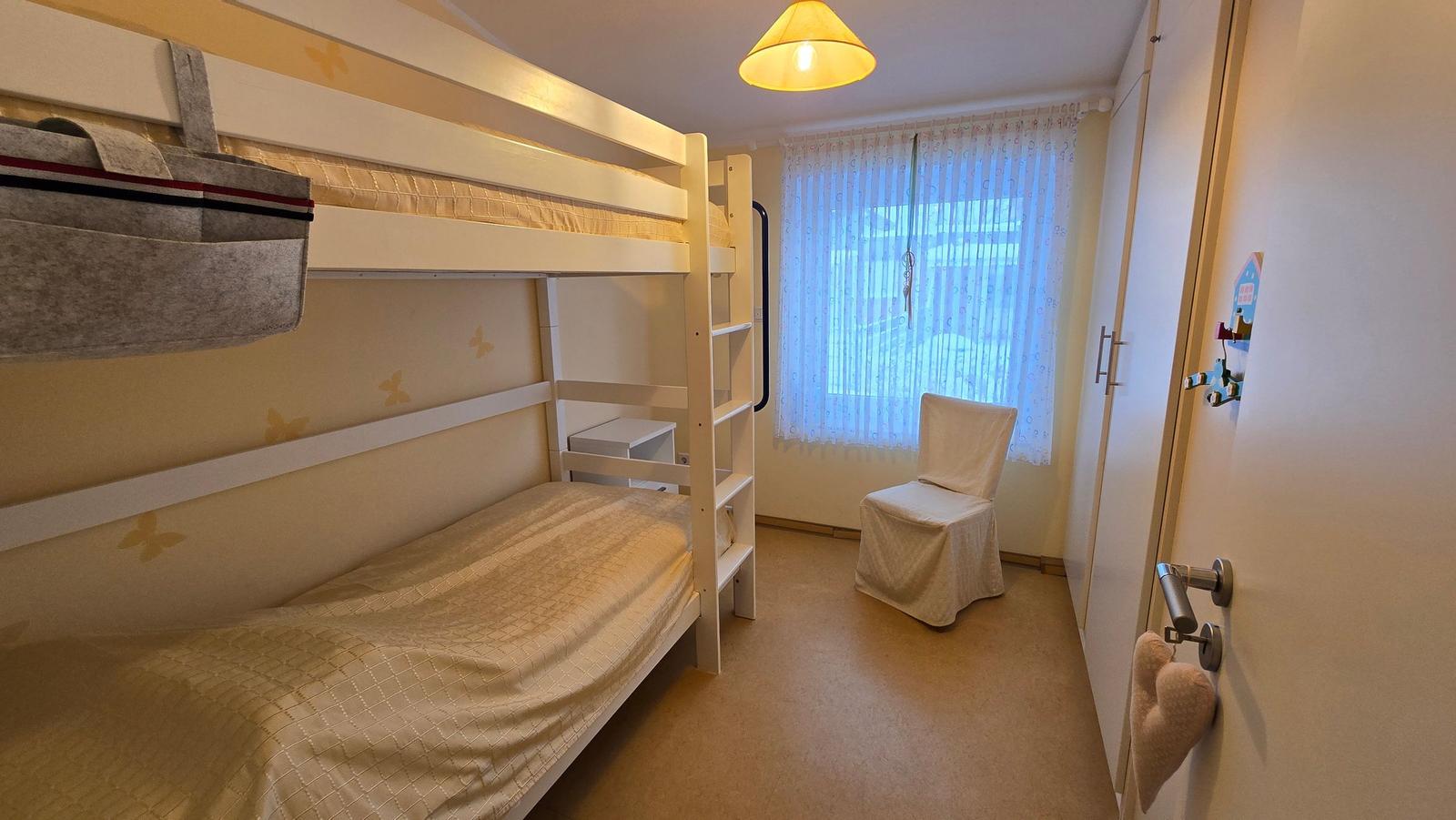 Kinderzimmer mit Doppelstockbett, Stuhl und Fenster mit Vorhängen.