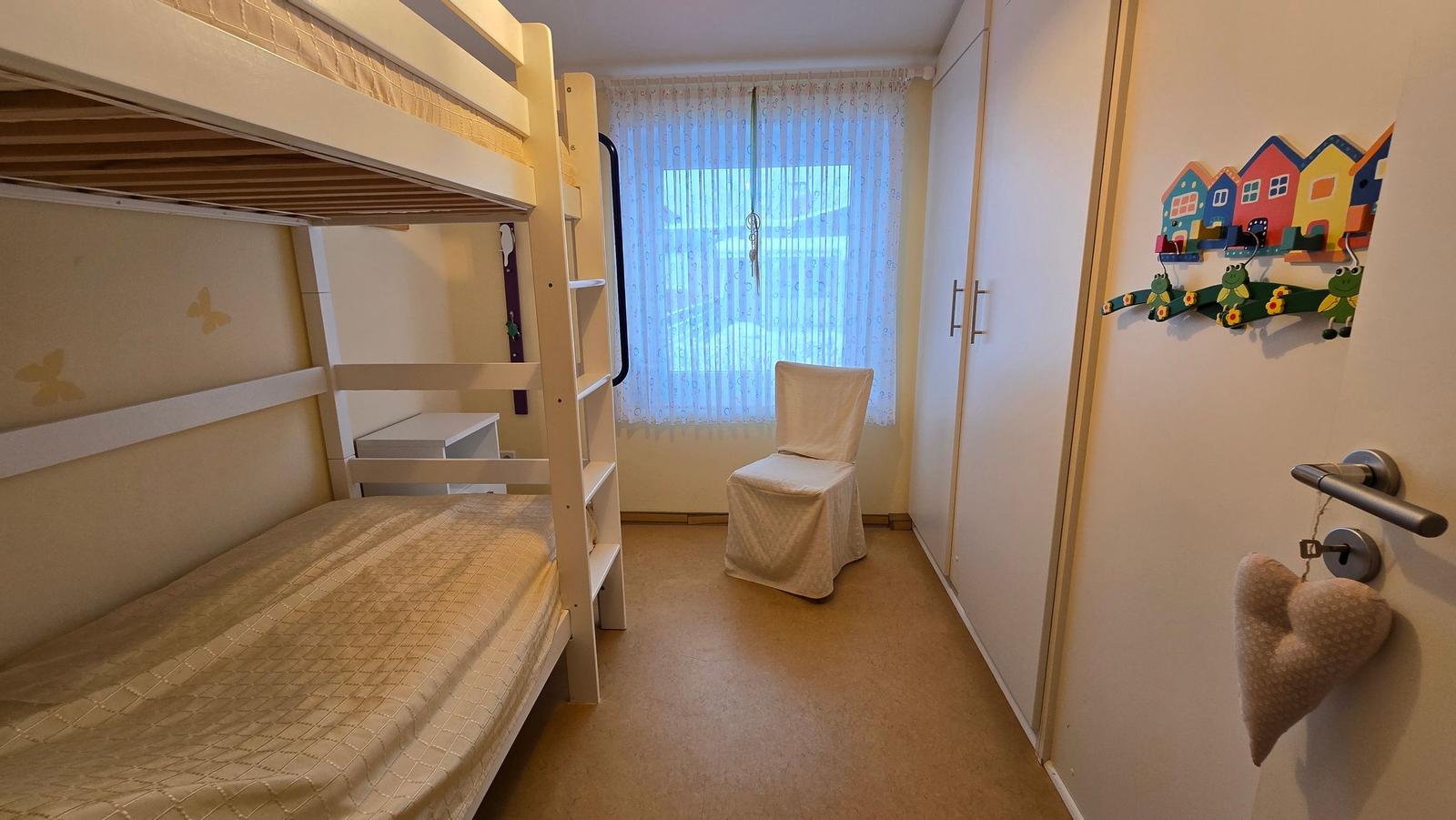 Zimmer mit Doppelstockbett, Stuhl und Kleiderschrank. Fenster mit Vorhängen.