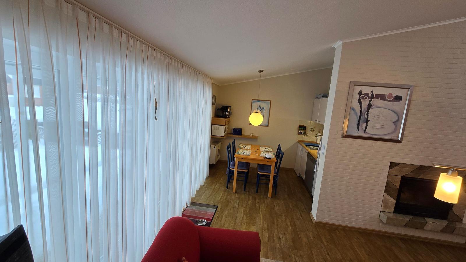 Wohnung mit Essbereich, Küche und Kaminofen. Durchgehende Holzdielen und weiße Vorhänge.