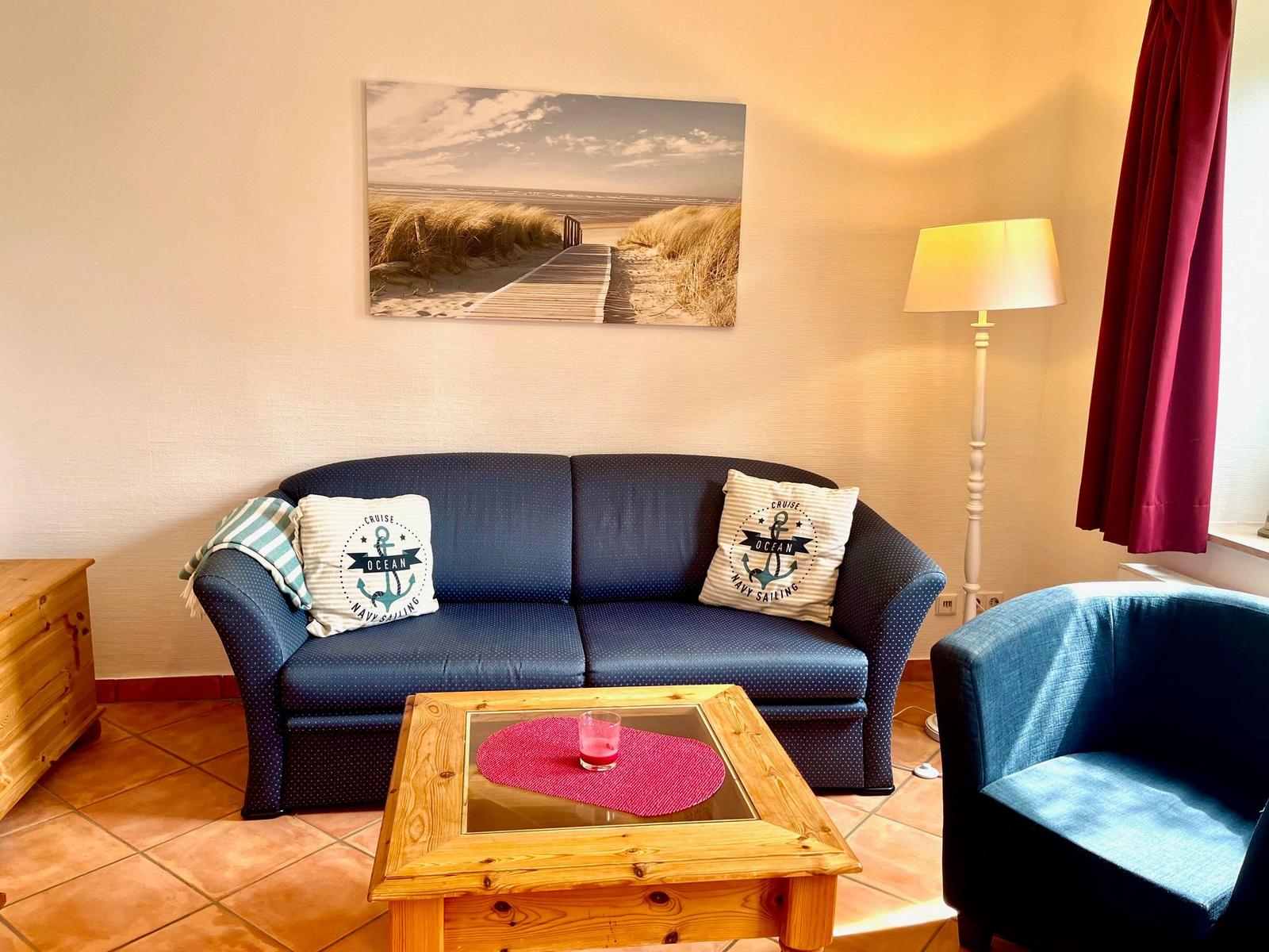 Ein Wohnzimmer mit Sofa, Sessel und Tisch. Wandbild zeigt Strand.
