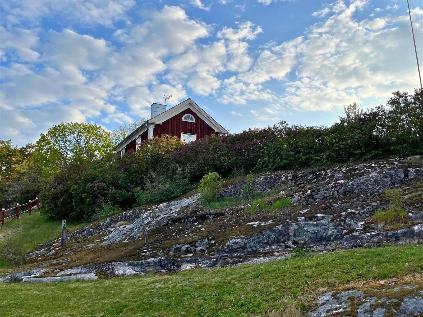 Rotes Haus auf grüner Anhöhe mit Steinhang und blauem Himmel.