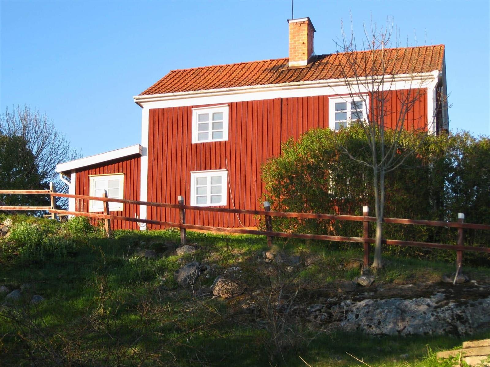 Rotes Holzhaus mit weißem Rahmen und Ziegeldach. Umgeben von Gras, Bäumen und einem Holzzaun.