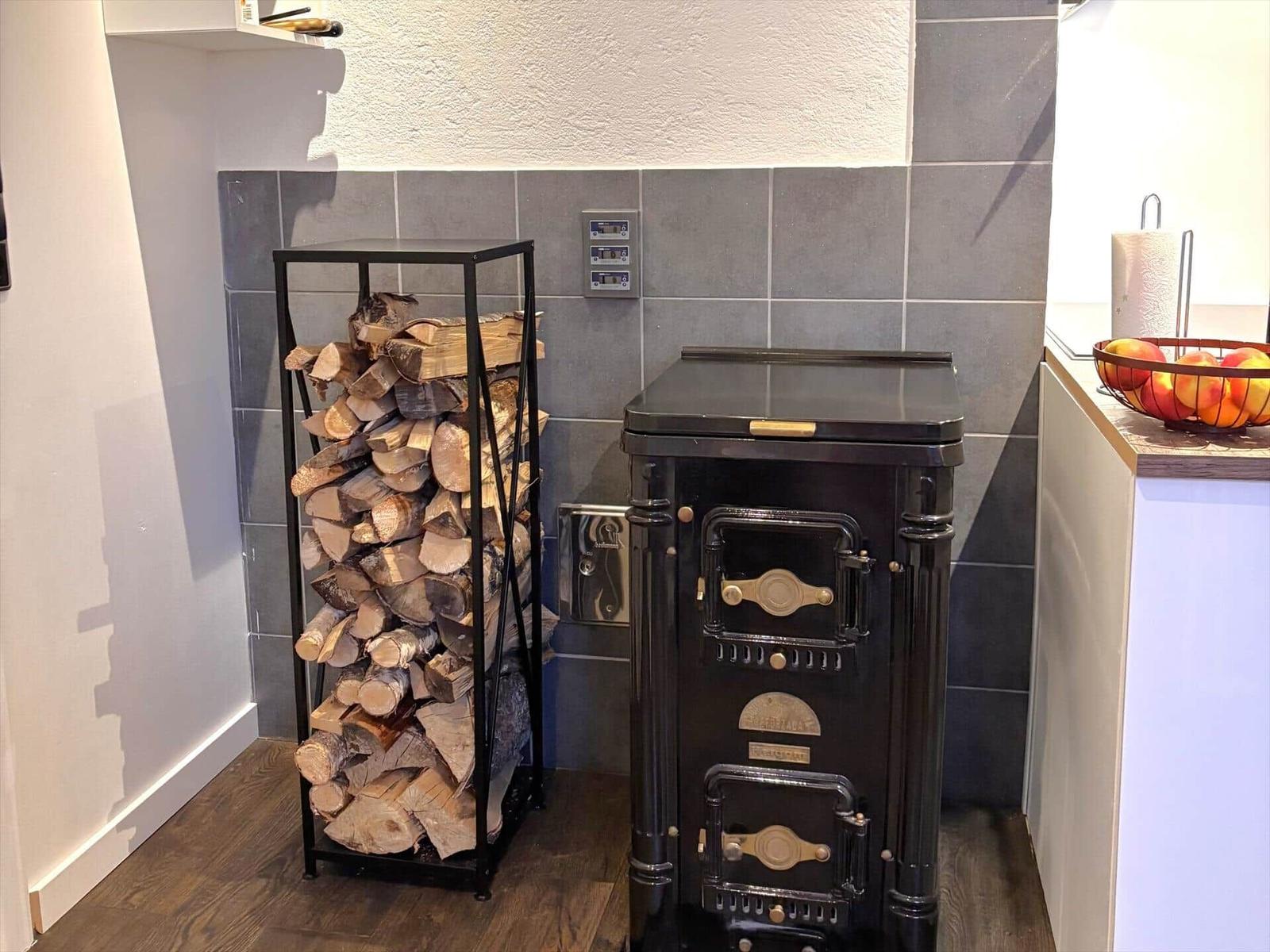 Schwarzer Ofen neben einem Holzstapel in einer Küche mit grauen Fliesen.