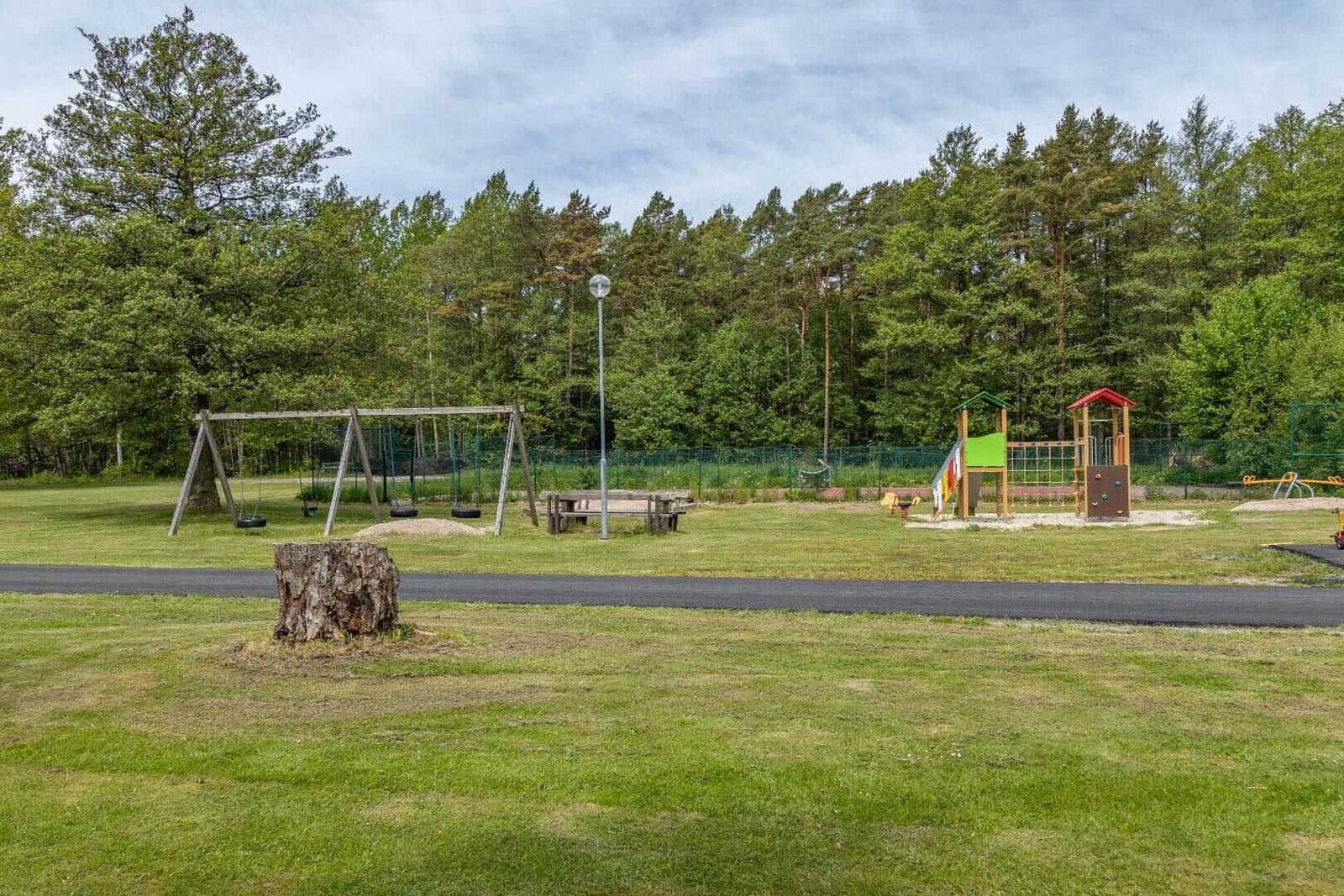 Grüner Spielplatz mit Schaukel, Klettergerüst und Holzbank vor Wald.