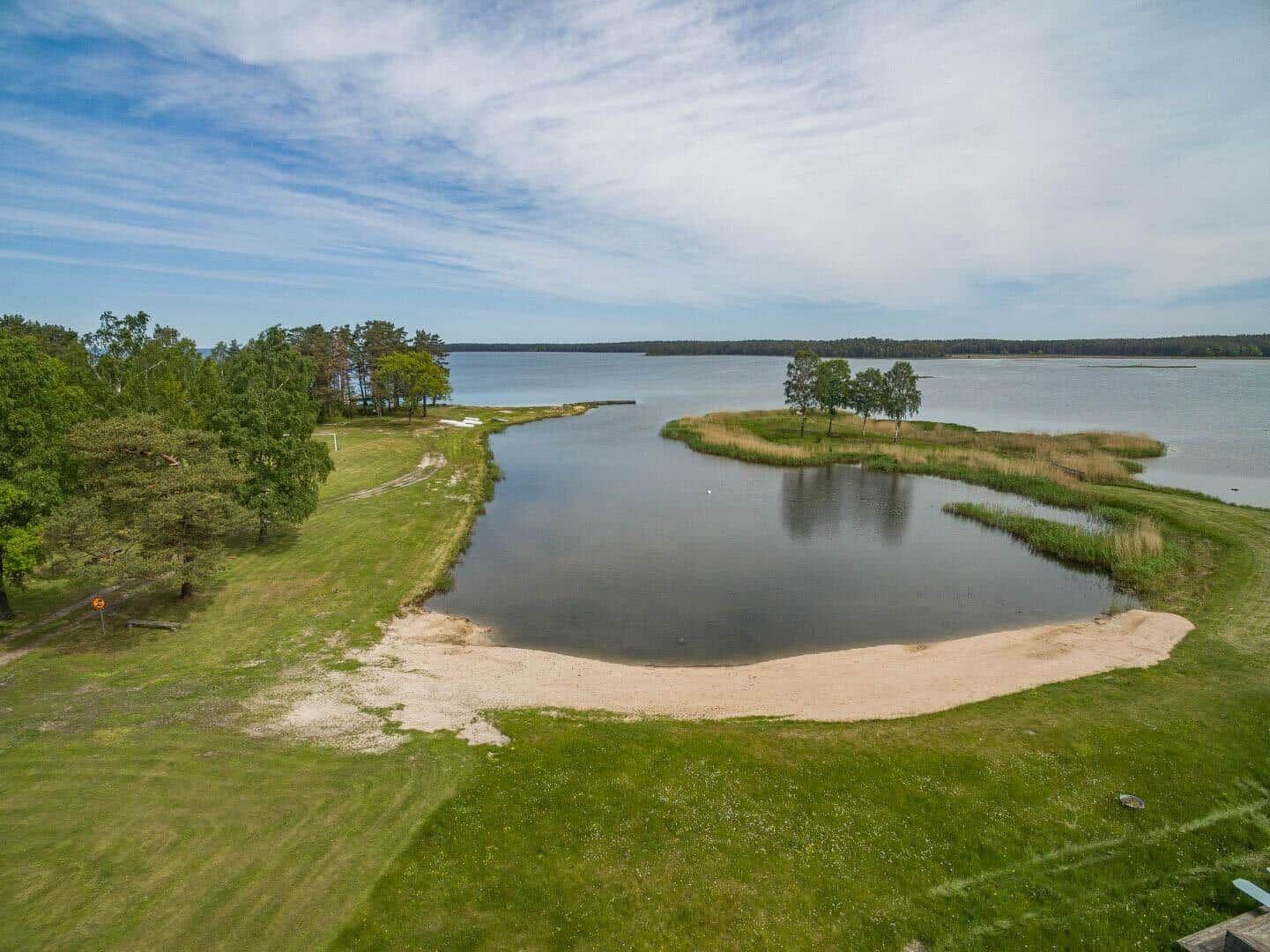 Landschaft mit See, Sandstrand und Grünflächen. Bäume und Insel im Hintergrund.