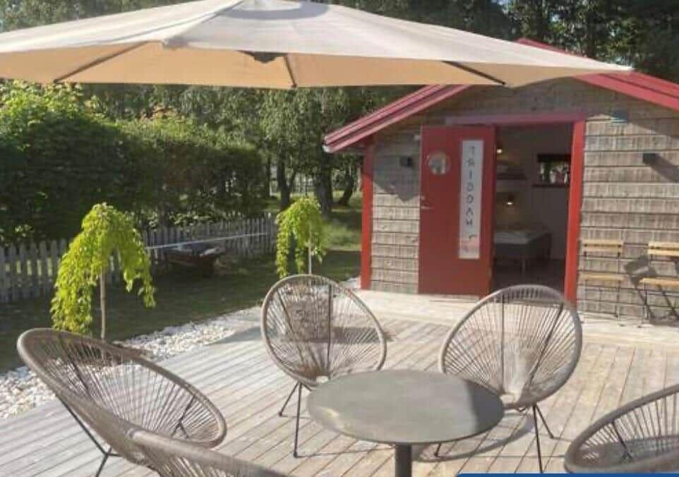 Terrasse mit Tisch und Stühlen unter einem Sonnenschirm. Hintergrund: Holzhaus mit roter Tür.