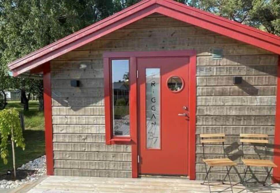 Rotes Holzhaus mit Tür und Fenster, zwei Stühle draußen.