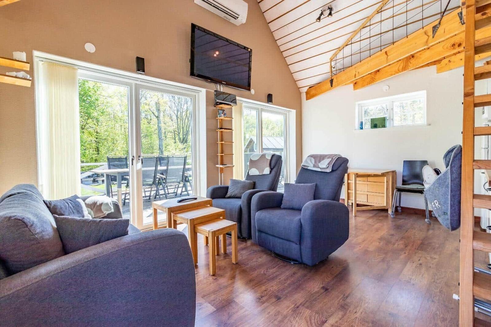 Wohnzimmer mit Sofa, zwei Sesseln und Holzboden. Große Fenster, TV und Treppe.