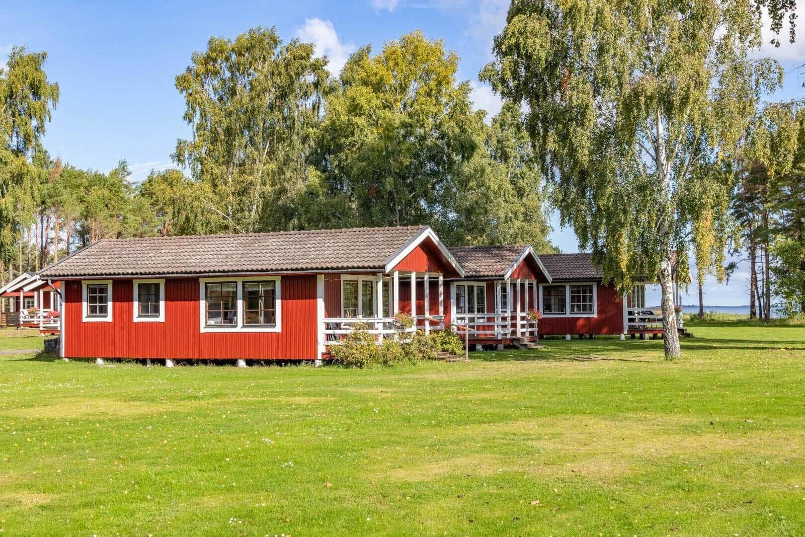 Rotes Ferienhaus mit weißem Balkon und grünem Rasen. Blick auf Wald und Wasser.