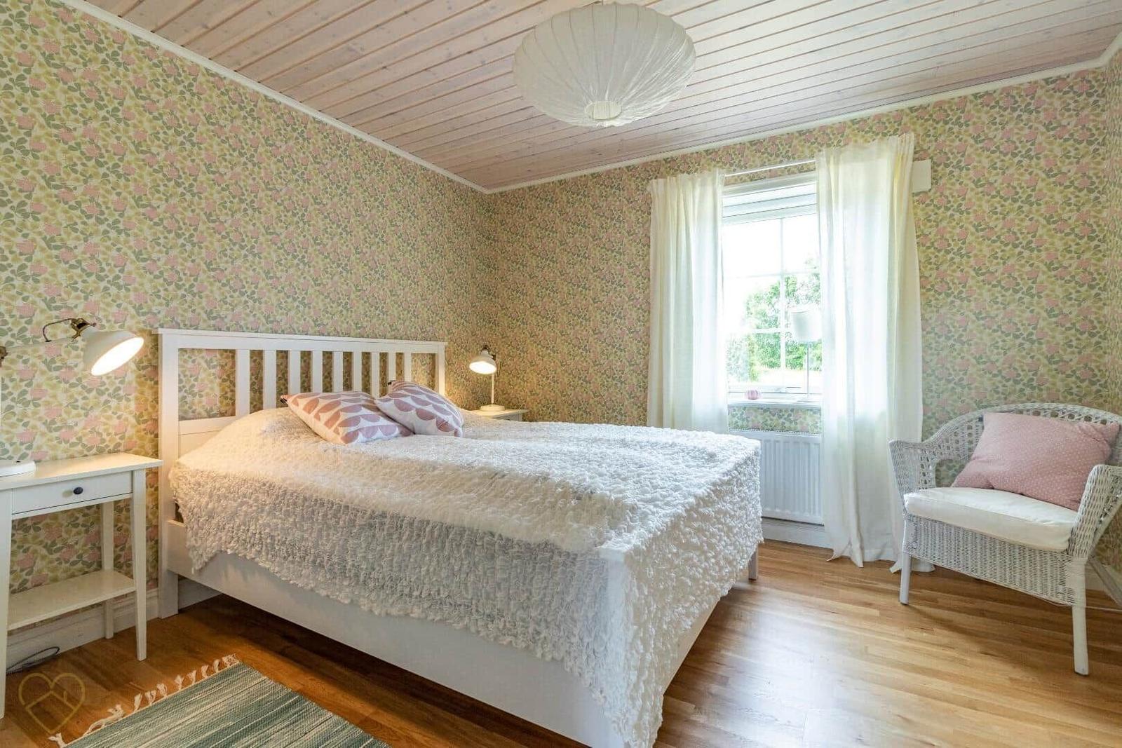 Schlafzimmer mit Bett, Stuhl und Fenster. Wände mit Tapete, Holzboden und Decke.