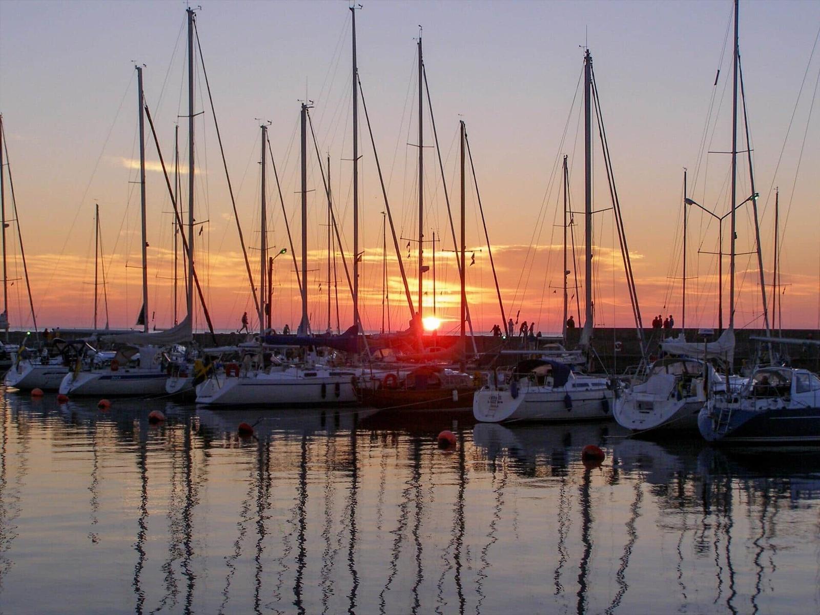 Sonnenuntergang über einem Hafen mit Segelyachten und Spiegelflächen.
