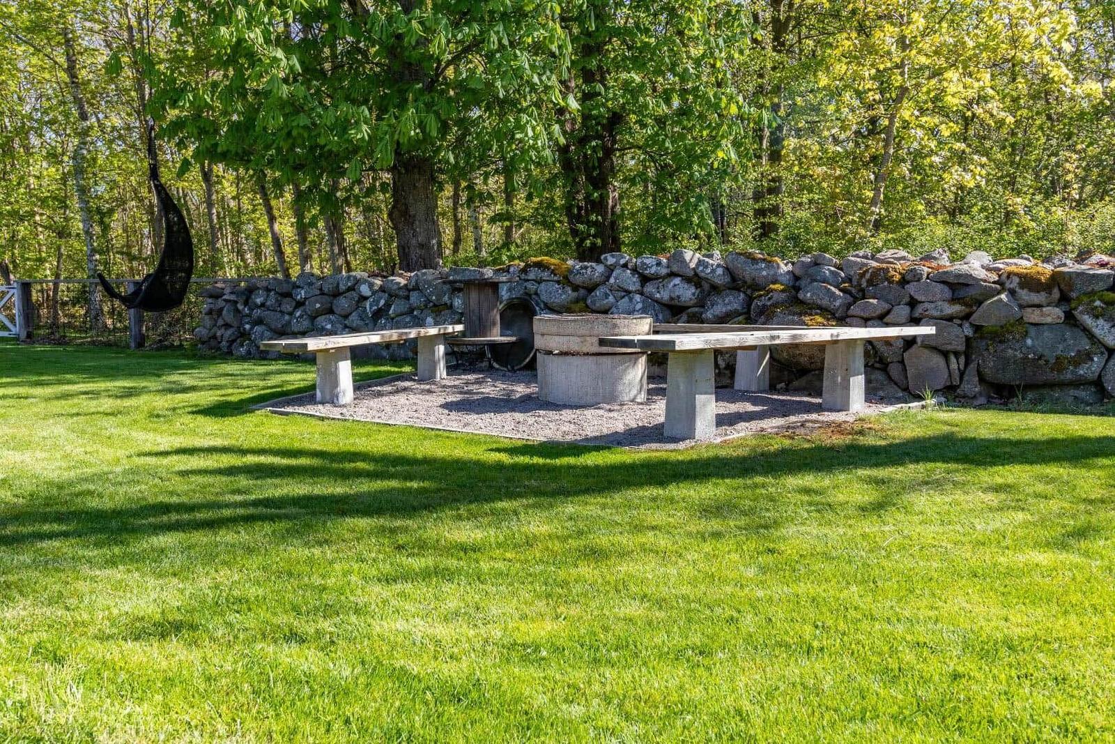 Hinterhof mit Steinmauer, Tisch, Bänken und Hängemöbel unter Bäumen.
