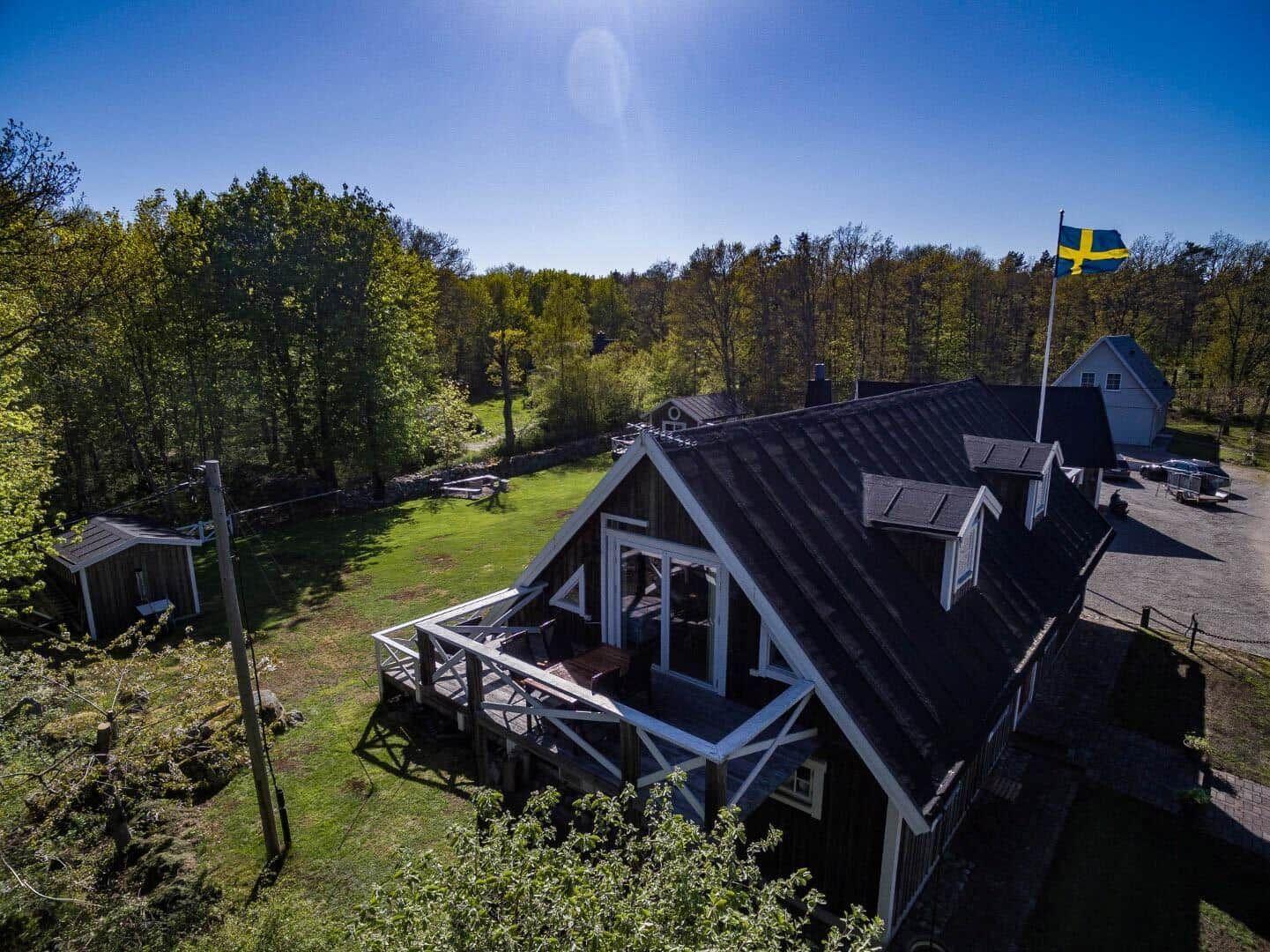 Holzhaus mit Terrasse, umgeben von Grün und Wald. Schwedische Flagge weht.