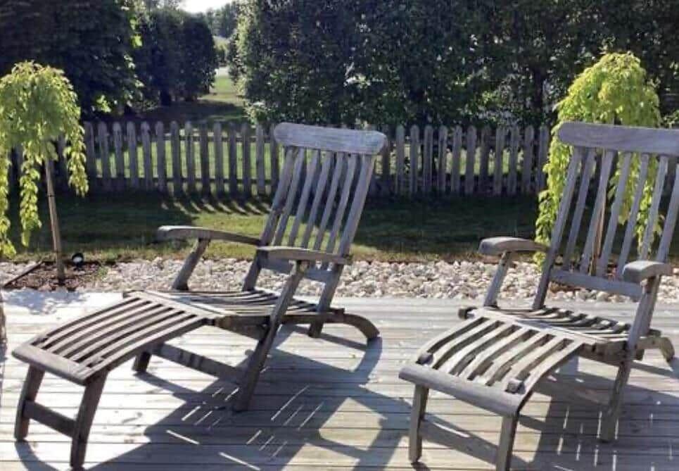 Zwei hölzerne Liegestühle auf einer Terrasse mit Grünfläche und Zaun im Hintergrund.