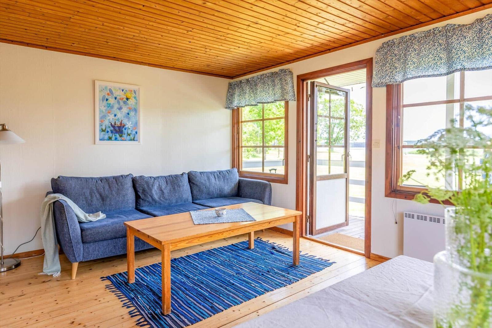 Wohnzimmer mit blauem Sofa, Holztisch und blauem Teppich. Holzdecke und Fenster mit Blick nach draußen.