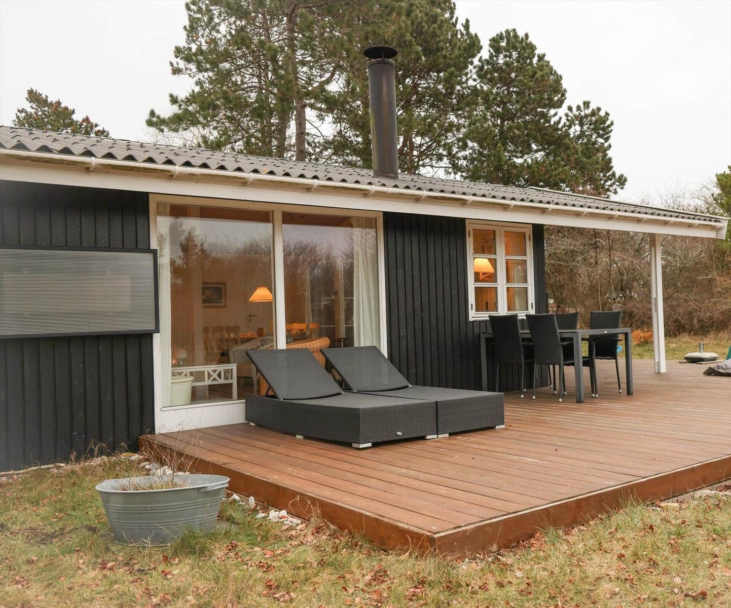 Holzterrasse mit Liegen und Tisch. Haus mit großen Fenstern und Schornstein.
