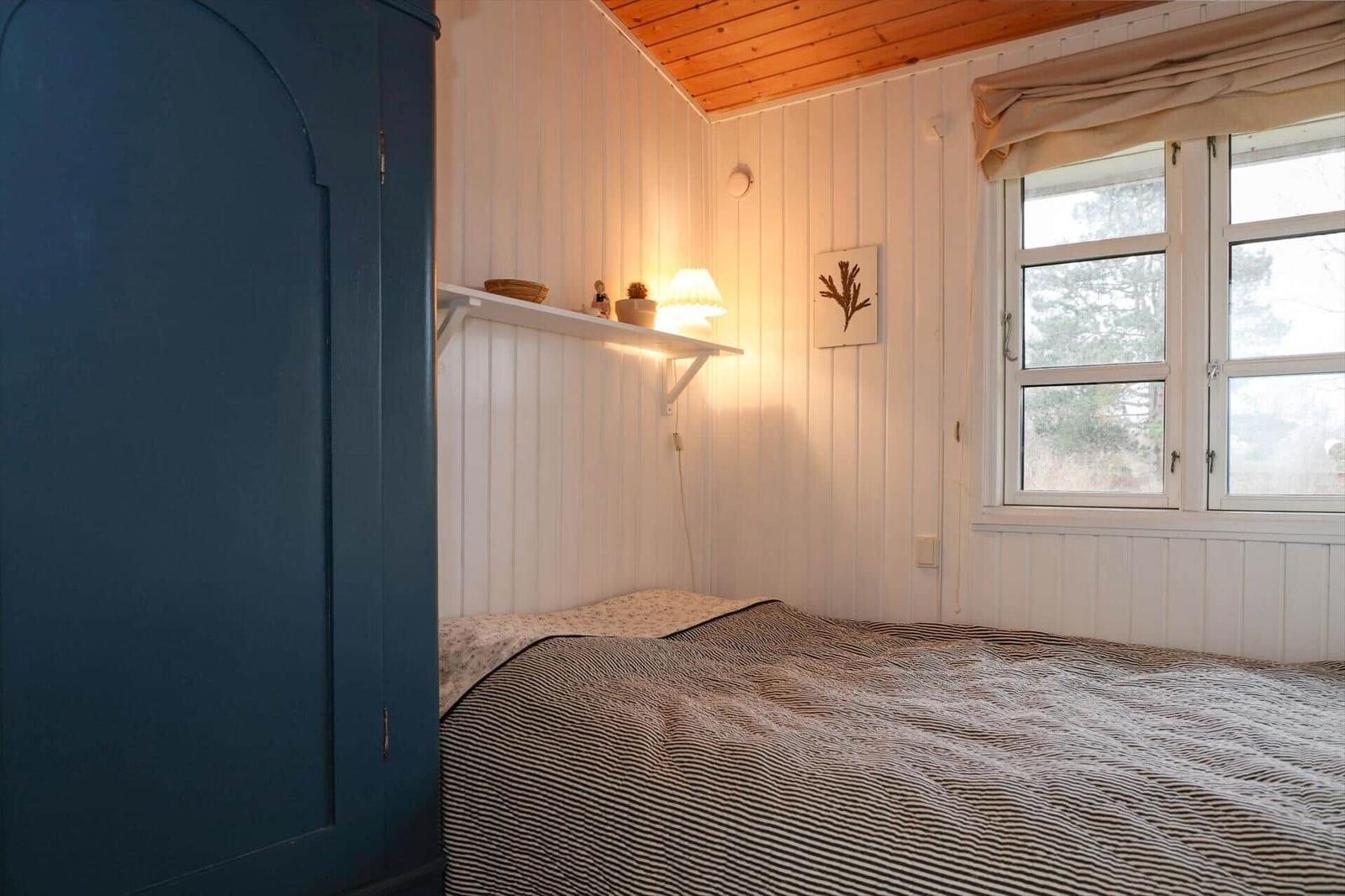 Schlafzimmer mit Bett, Kleiderschrank und Fenster. Wände mit weißen Paneelen, Holzdecke.