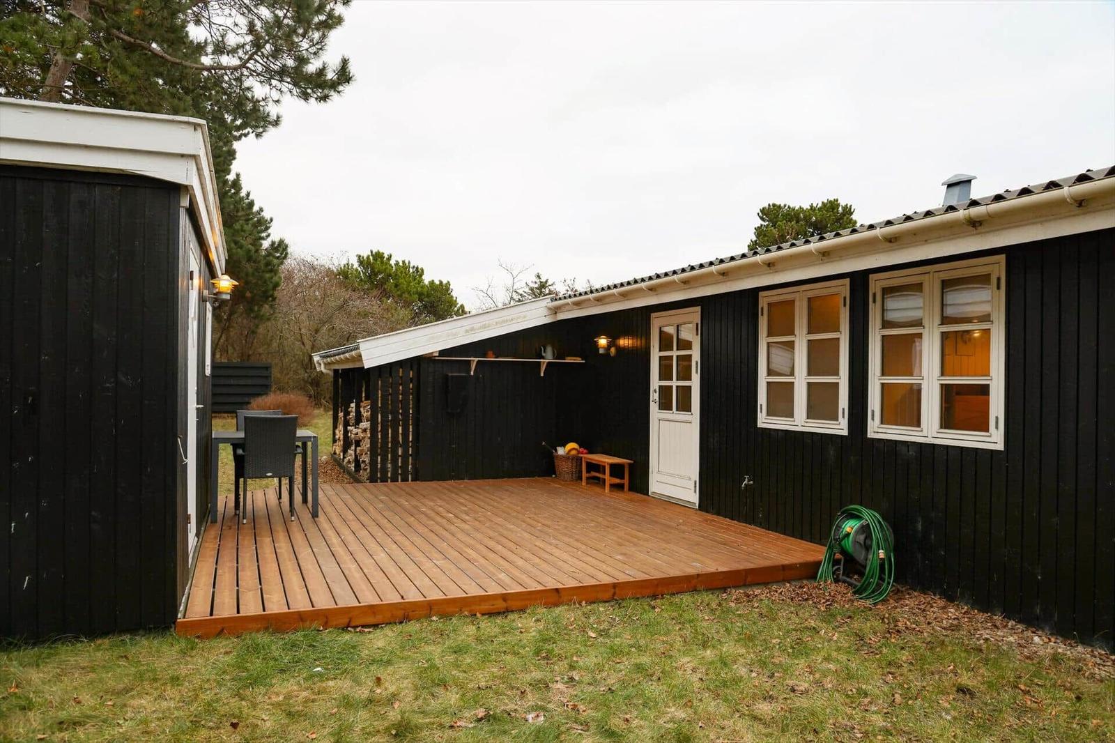 Holzterrasse mit Tisch und Stühlen vor einem schwarz getäfelten Haus mit weißen Fenstern.